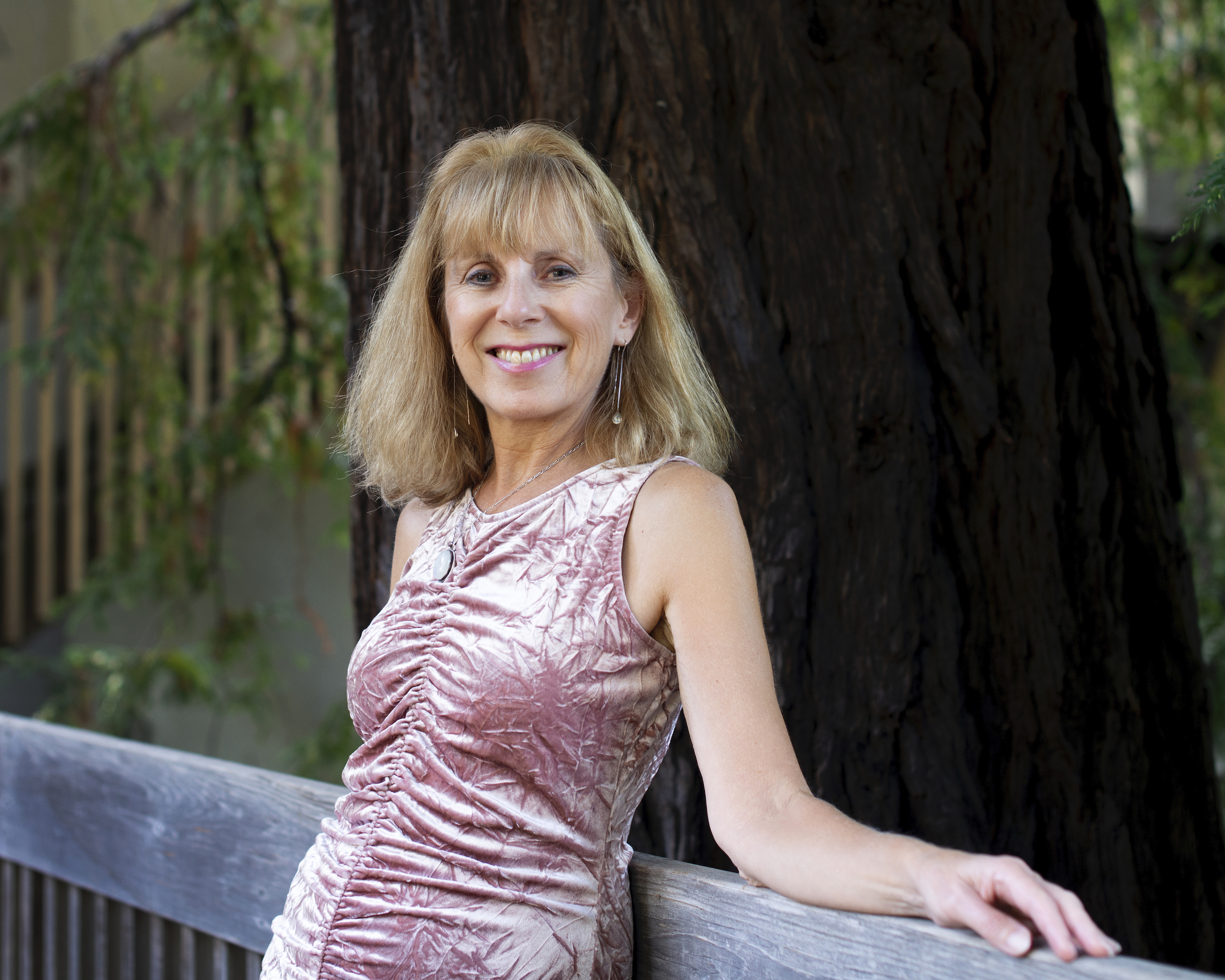 Nicola Walker in pink velvet dress leaning against railing in front of redwood tree