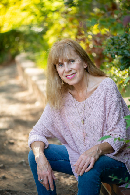 Nicola Walker wearing pale pink sweater and blue jeans sitting on low wall with green foliage