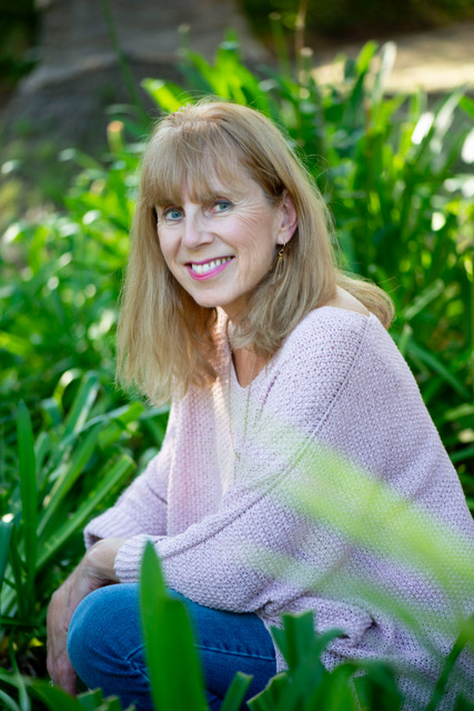 Nicola Walker wearing pale pink sweater and blue jeans with green foliage in background